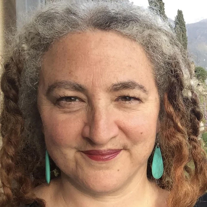 Image of an Arab Jewish woman with jewelry and a gray and black striped top set in front of a dark wooden background